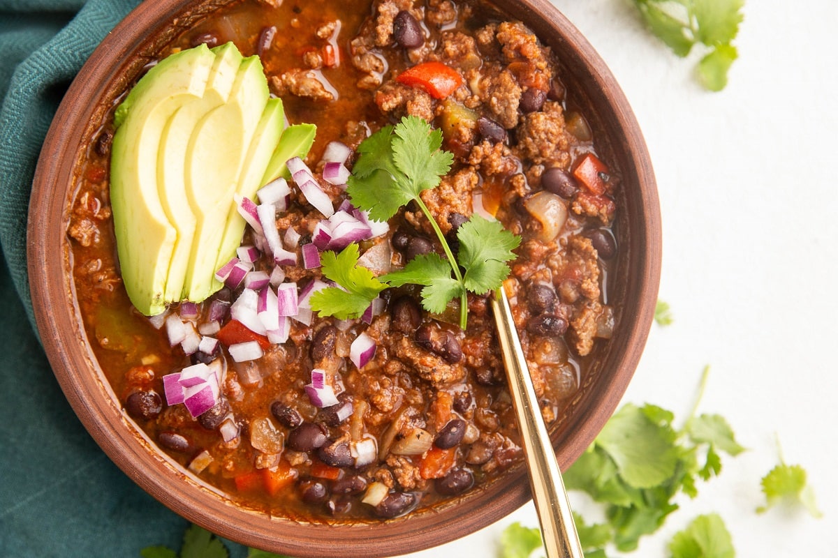 Big brown bowl of chili with avocado, onion, and cilantro on top, ready to eat.