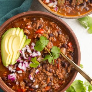 Two bowls of beef chili with avocado, red onion and cilantro on top. A blue napkin to the side.