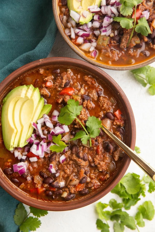 Two bowls of beef chili with avocado, red onion and cilantro on top. A blue napkin to the side.