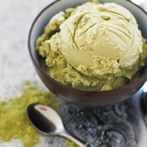 Scoop of green tea ice cream in a bowl with a spoon to the side and a sprinkle of matcha powder to the side.