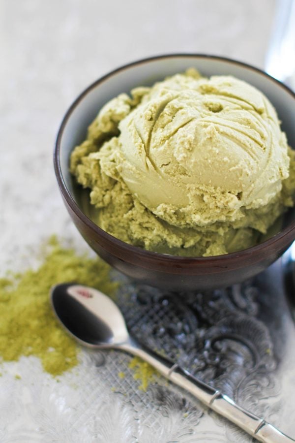 Scoop of green tea ice cream in a bowl with a spoon to the side and a sprinkle of matcha powder to the side.