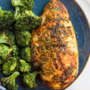 Blue plate with cooked chicken breast and broccoli on top with a red napkin to the side.