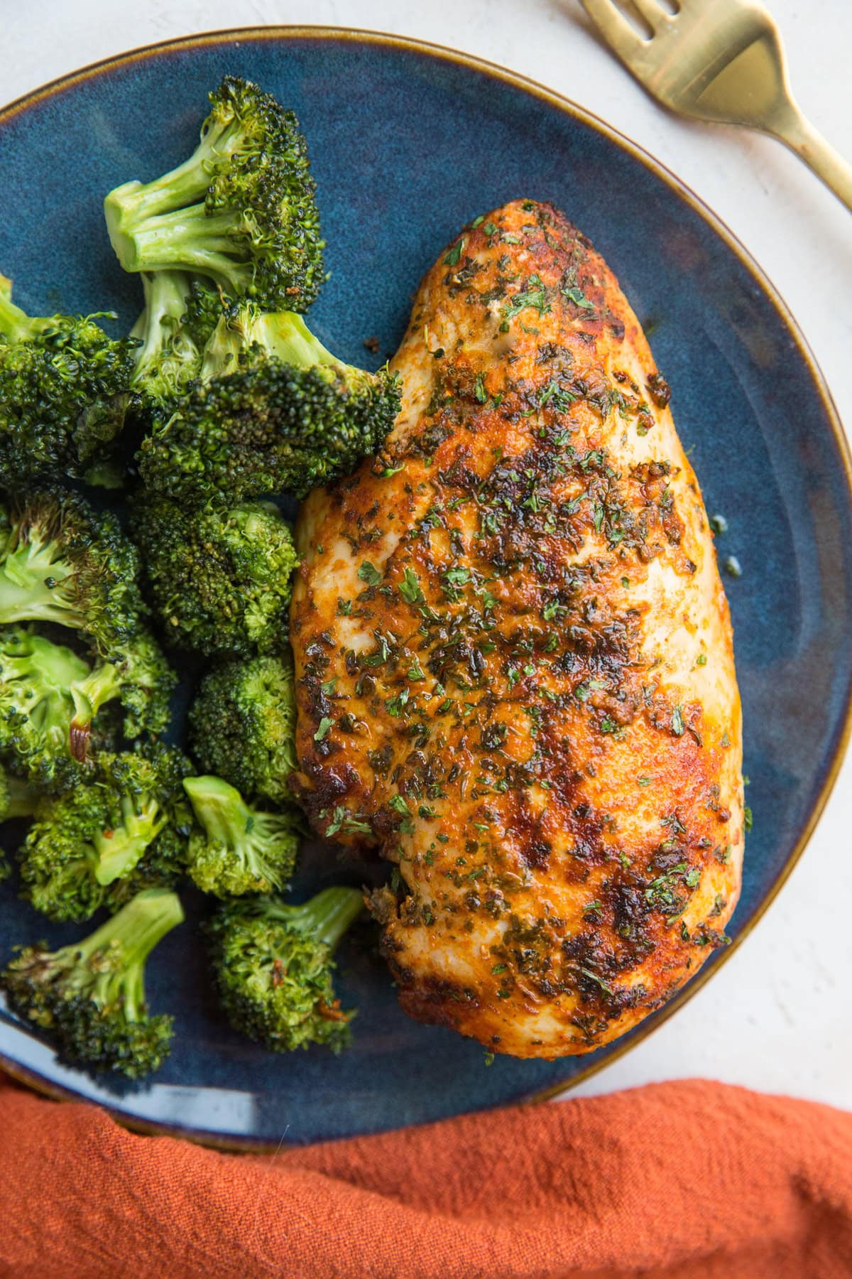 Blue plate with cooked chicken breast and broccoli on top with a red napkin to the side.