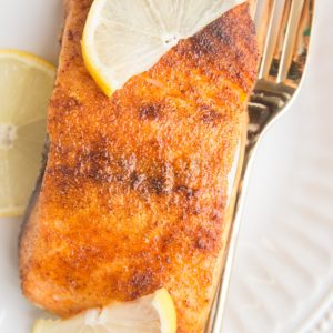 Crispy salmon on a plate with slices of lemon and a golden fork. A close up image of cooked salmon where you can see the salmon filet glistening.