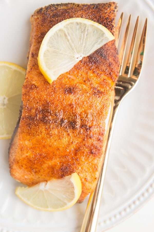 Crispy salmon on a plate with slices of lemon and a golden fork. A close up image of cooked salmon where you can see the salmon filet glistening.