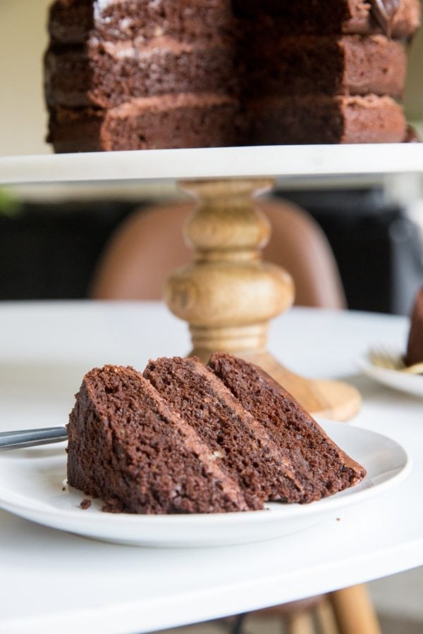 Slice of chocolate cake on a white plate with another plate of cake in the background and the cake stand with the rest of the cake in the background. Ready to serve.