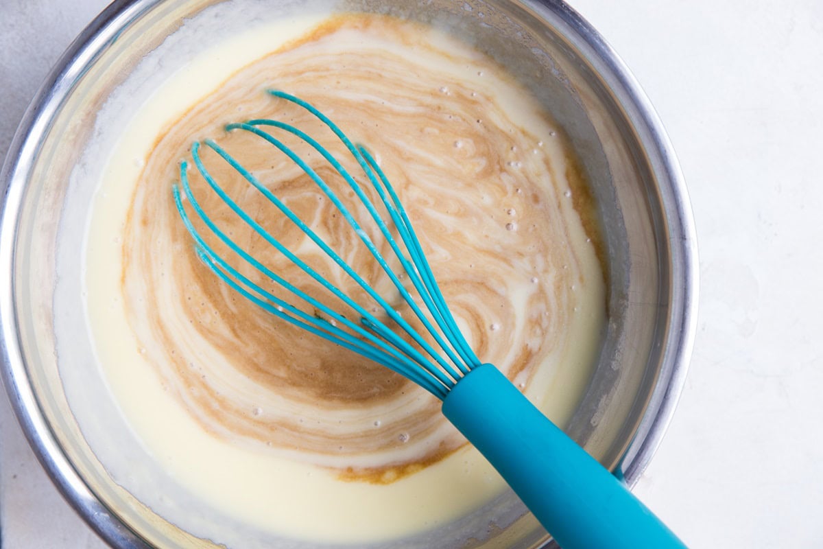 Coconut milk, coffee, eggs, and oil being mixed together in a stainless steel mixing bowl with a whisk.