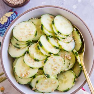 White ceramic bowl with cucumber salad inside, sprinkled with sesame seeds. A golden spoon for serving.