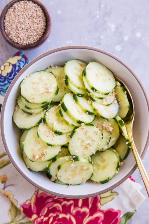 White ceramic bowl with cucumber salad inside, sprinkled with sesame seeds. A golden spoon for serving.