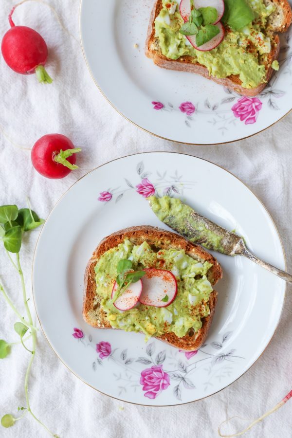 Two plates with two avocado egg salad toasts on top. Fresh microgreens and radishes to the side.