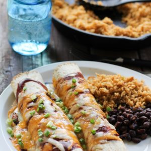 BBQ Pulled Pork Enchiladas covered with cheese and bbq sauce on a plate with side dishes and a glass of water and skillet of rice in the background.