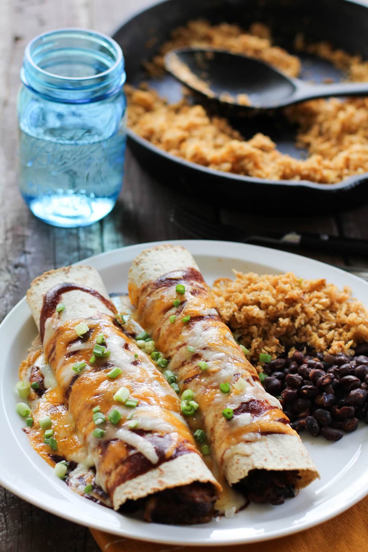 BBQ Pulled Pork Enchiladas covered with cheese and bbq sauce on a plate with side dishes and a glass of water and skillet of rice in the background.