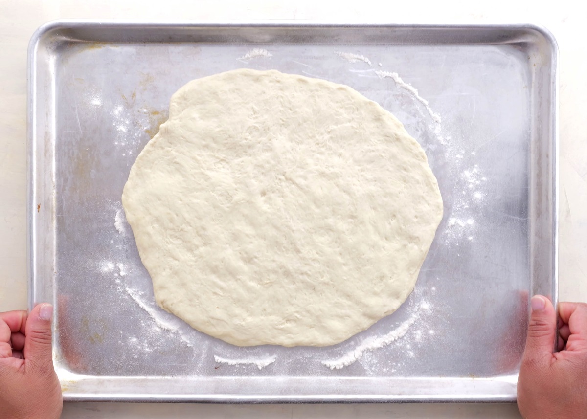 Pizza dough spread out onto a baking sheet with flour beneath it, ready to go into the oven to prebake.