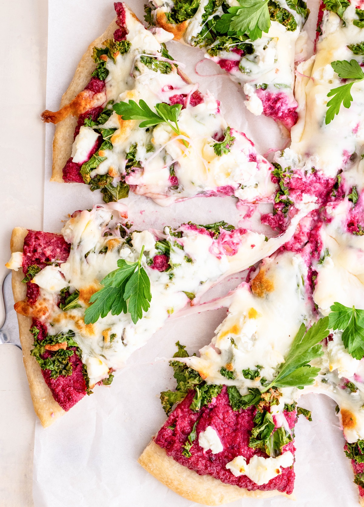 Pizza cut into slices on a sheet of parchment paper with beet pesto sauce, kale, and goat cheese.