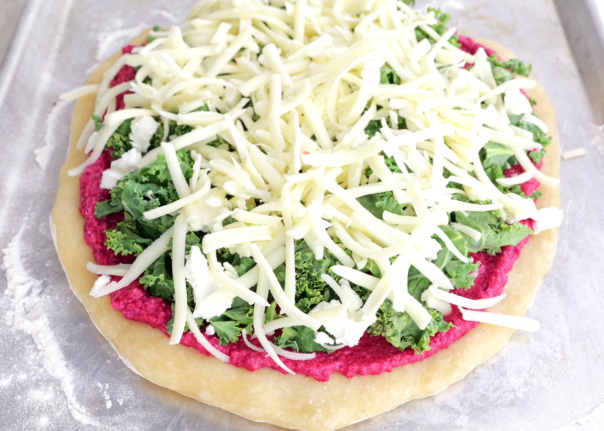 Pizza crust with beet pesto sauce, chopped kale, goat cheese, and mozzarella cheese on top, ready to go into the oven.