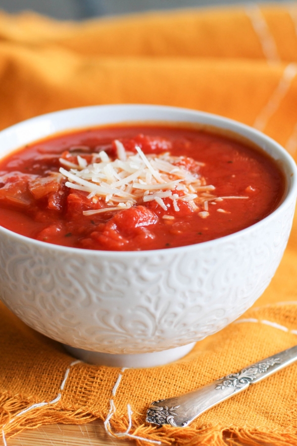 Small white bowl of chunky tomato soup with parmesan cheese on top and an orange napkin and silver spoon.