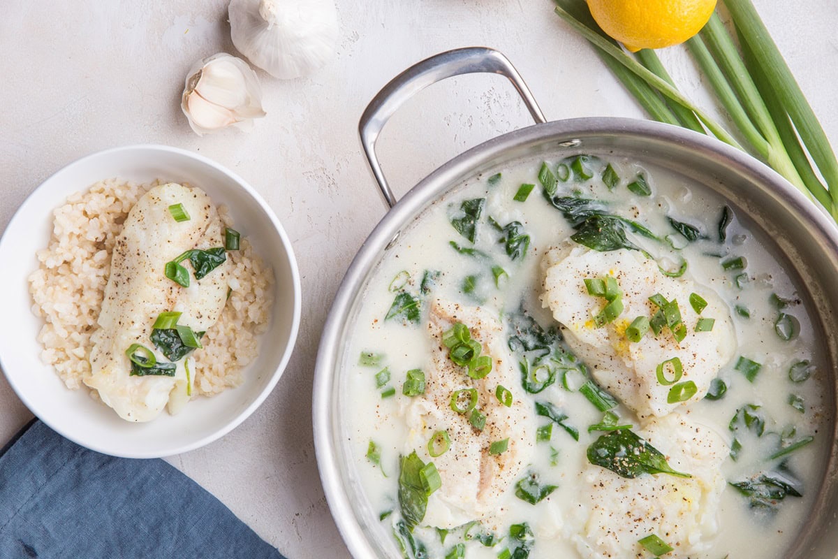 white bowl of brown rice and a cod filet with a skillet of cod fillets in a creamy lemon sauce.