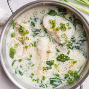 Creamy Lemon Garlic Cod recipe in a stainless steel skillet with a blue napkin to the side and fresh green onion and garlic next to the skillet.