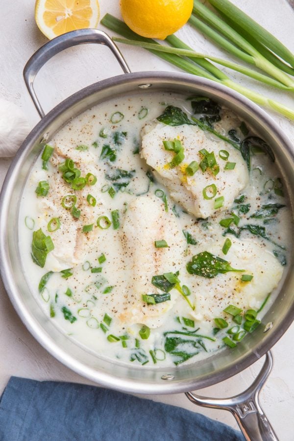 Creamy Lemon Garlic Cod recipe in a stainless steel skillet with a blue napkin to the side and fresh green onion and garlic next to the skillet.