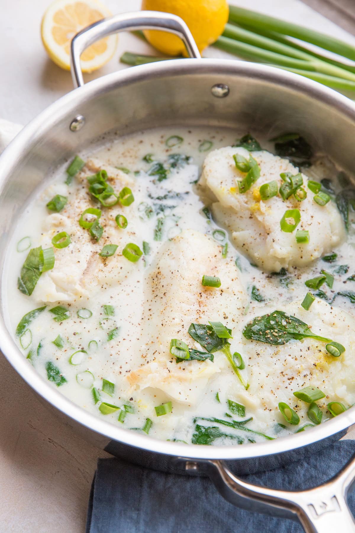 Cod fillets in a stainless steel skillet with a creamy lemon garlic sauce, sprinkled with fresh green onions.