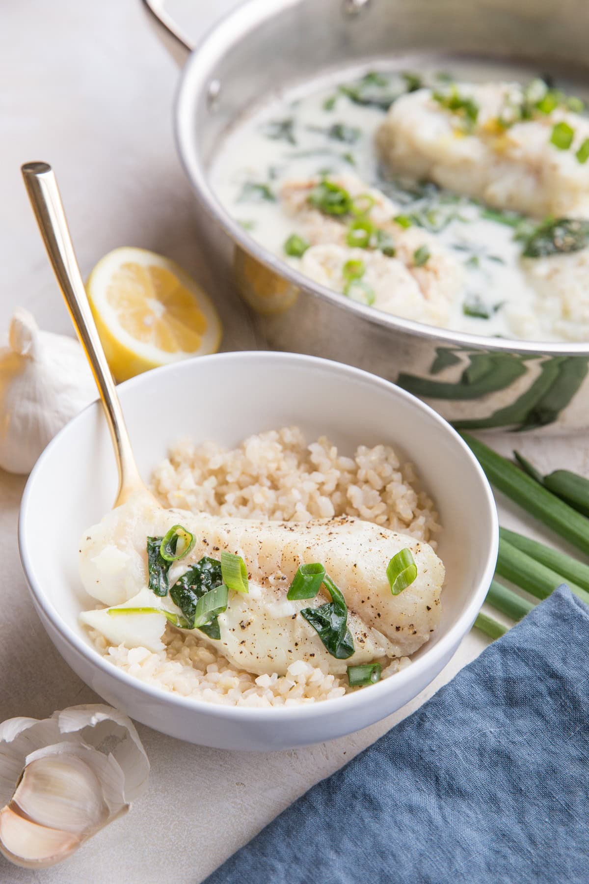 White bowl of rice and a cod fish filet on top with fresh garlic and a blue napkin to the side and the skillet of the rest of the fish fillets in the background.