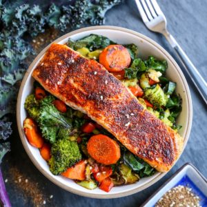 Bowl of crispy salmon on top of roasted vegetables with a fresh kale leaf to the side.