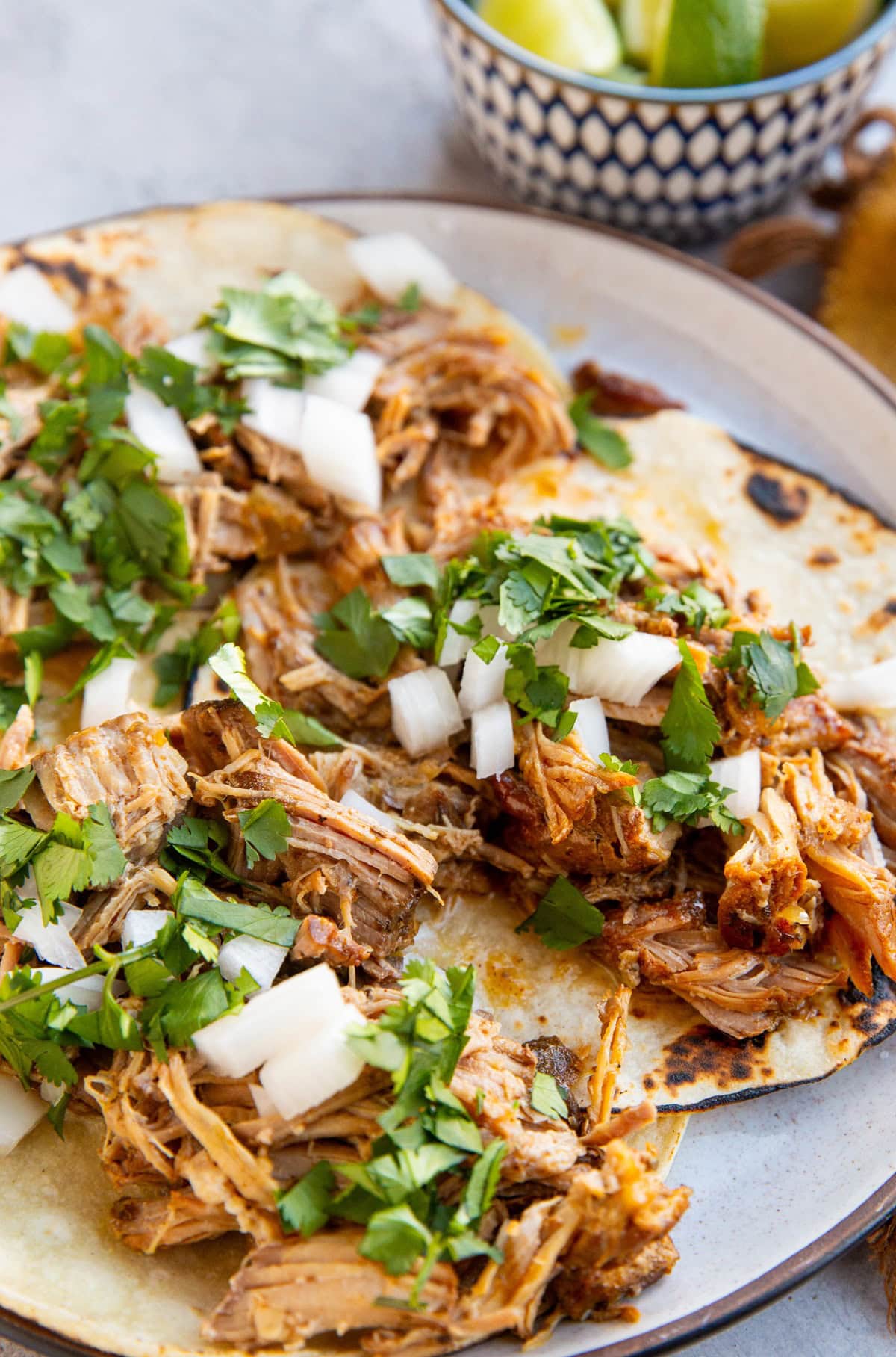 Plate with three carnitas tacos and a small bowl of lime wedges in the background.