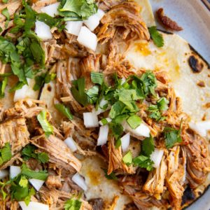 Shredded pork tacos on a plate with cilantro and white onion sprinkled on top.