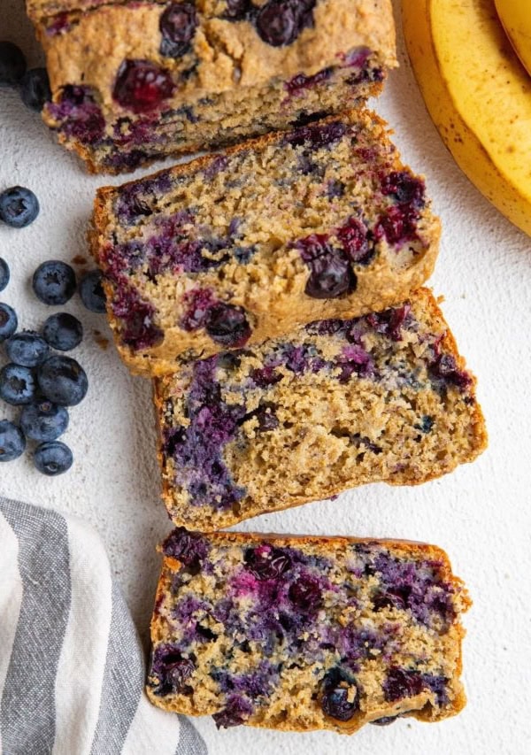 Loaf of banana oatmeal bread cut into slices with ripe bananas and blueberries to the side.