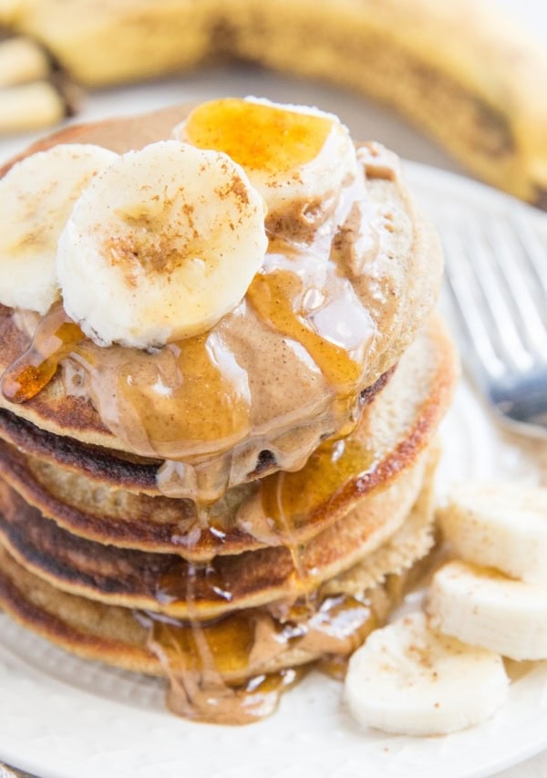 Stack of oatmeal banana pancakes on a plate with almond butter, honey, and sliced bananas on top.