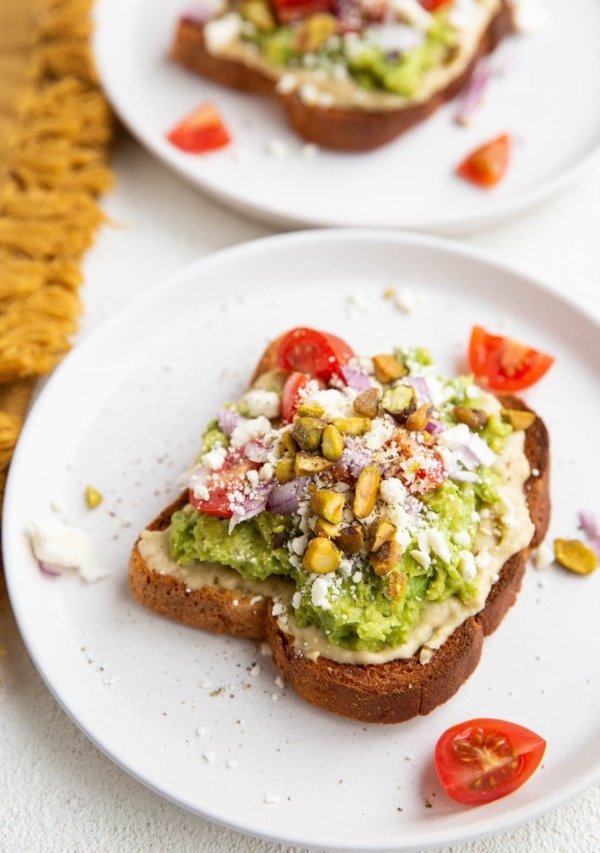 Two slices of avocado hummus toast on white plates, ready to eat.