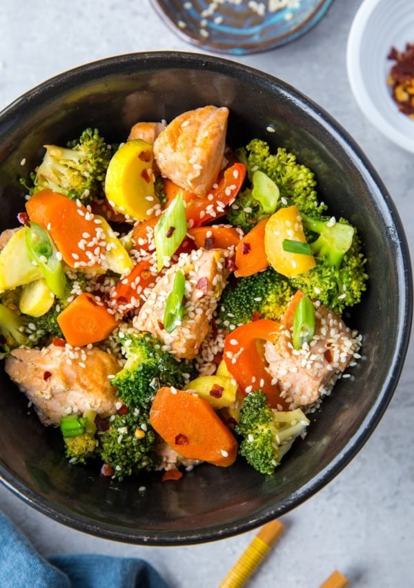 Black bowl of stir fry salmon and vegetables with chopsticks to the side and a dark blue napkin, ready to consume.