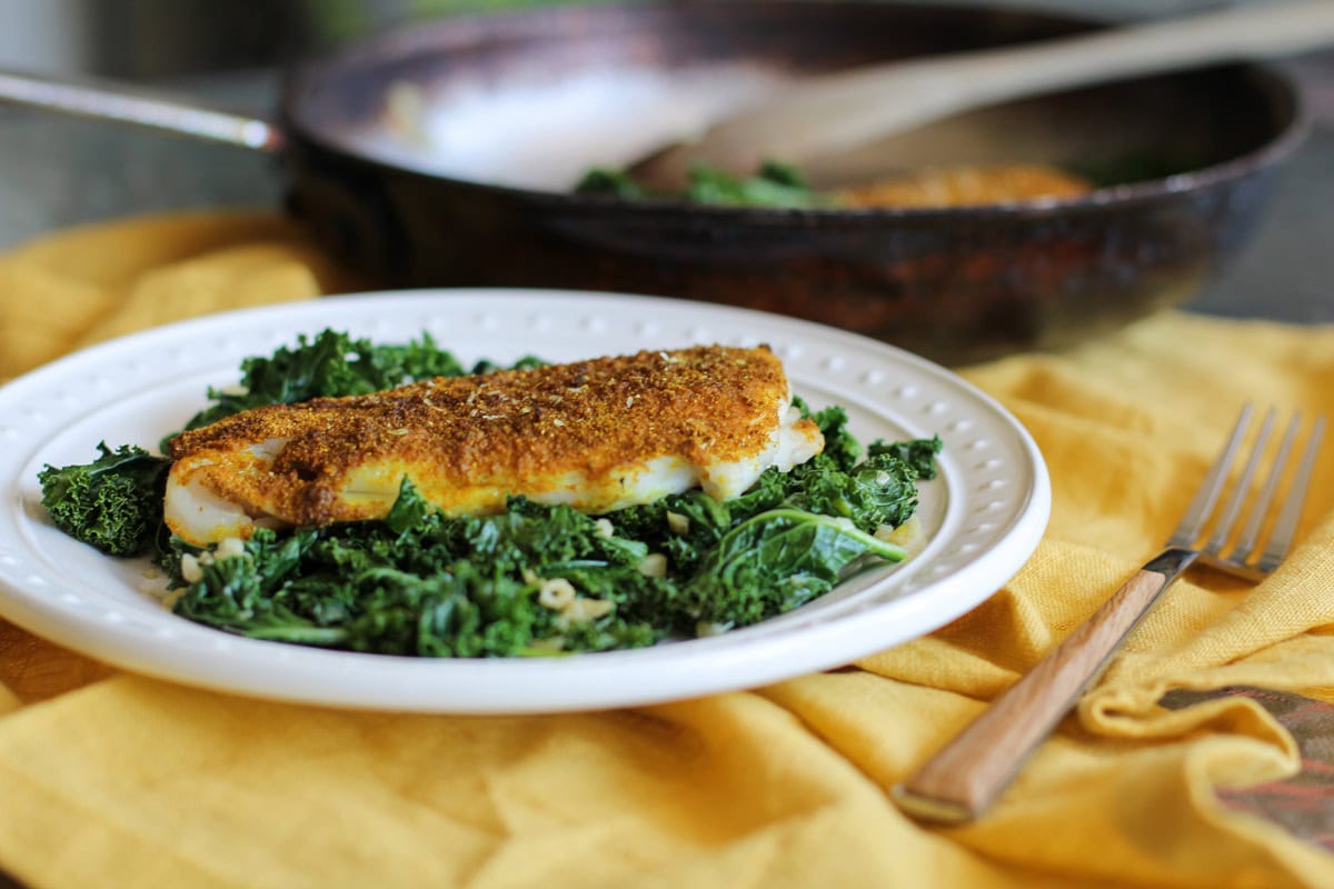 White plate of curry sole on top of kale with a yellow napkin and a fork to the side.