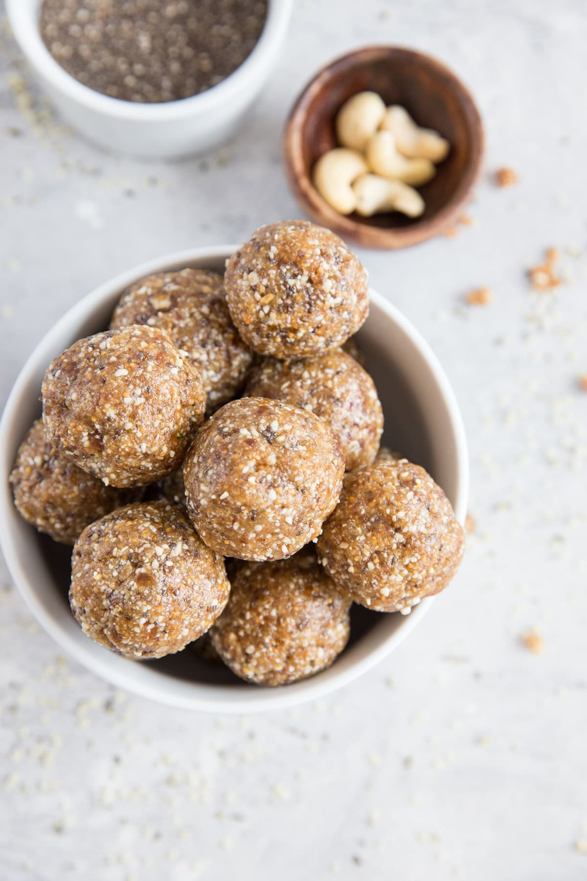 Bowl of fig and date energy balls with a bowl of cashews and a bowl of chia seeds to the side and sprinkles of hemp seeds all around.