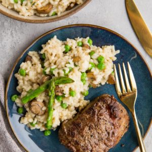 Filet mignon on a blue plate with mushroom risotto and a gold fork, ready to eat.