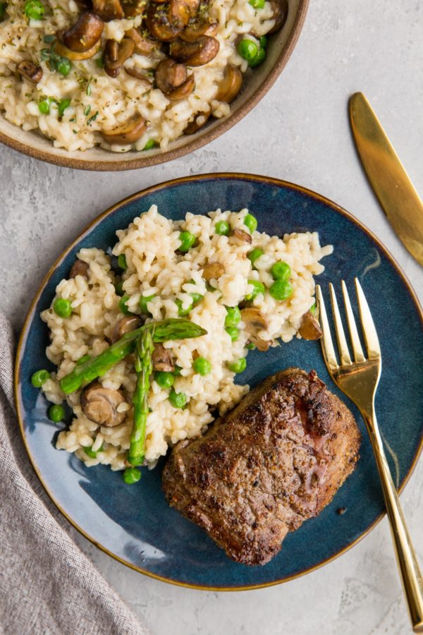 Filet mignon on a blue plate with mushroom risotto and a gold fork, ready to eat.