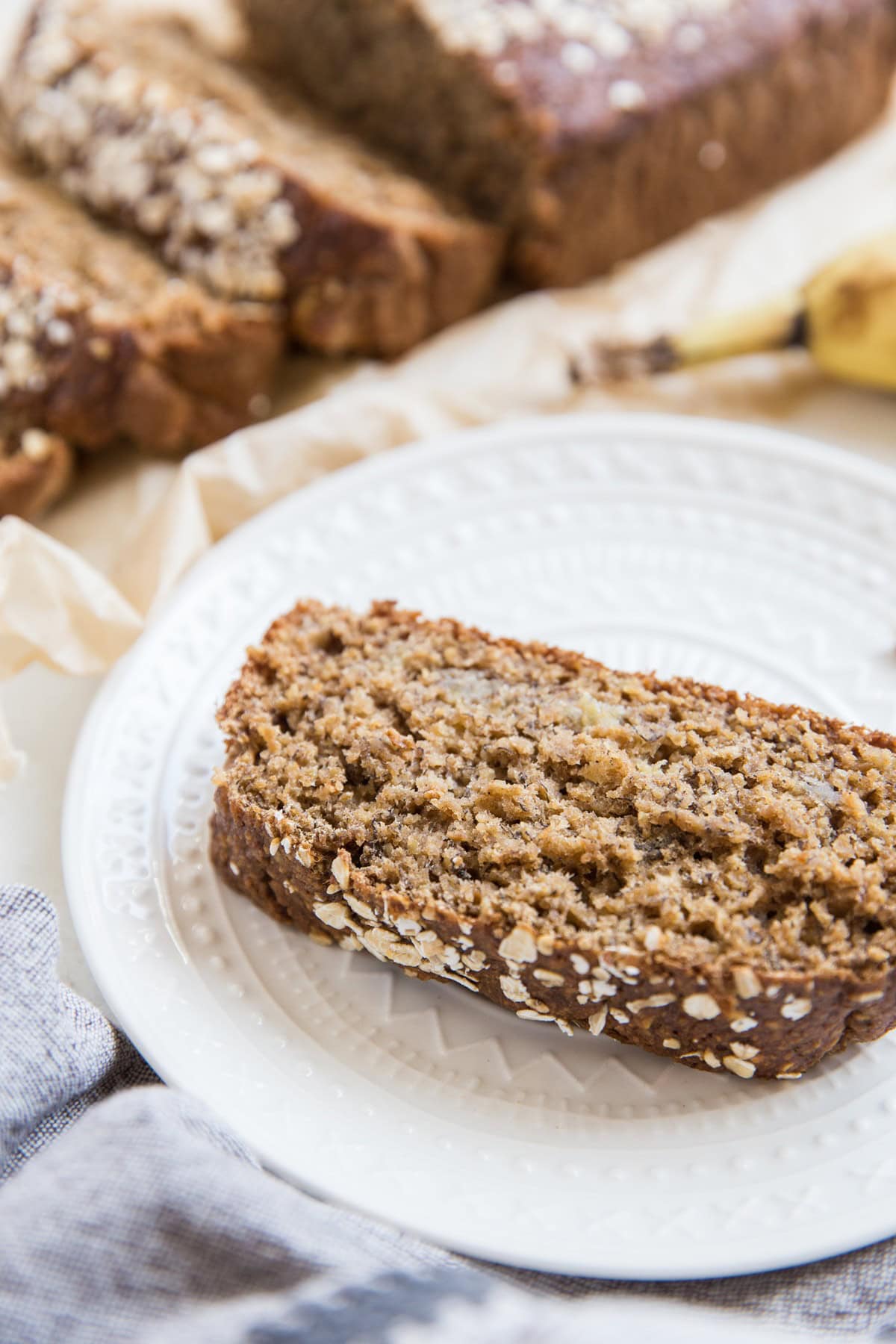 Slice of banana bread on a white plate with the rest of the loaf in the background.