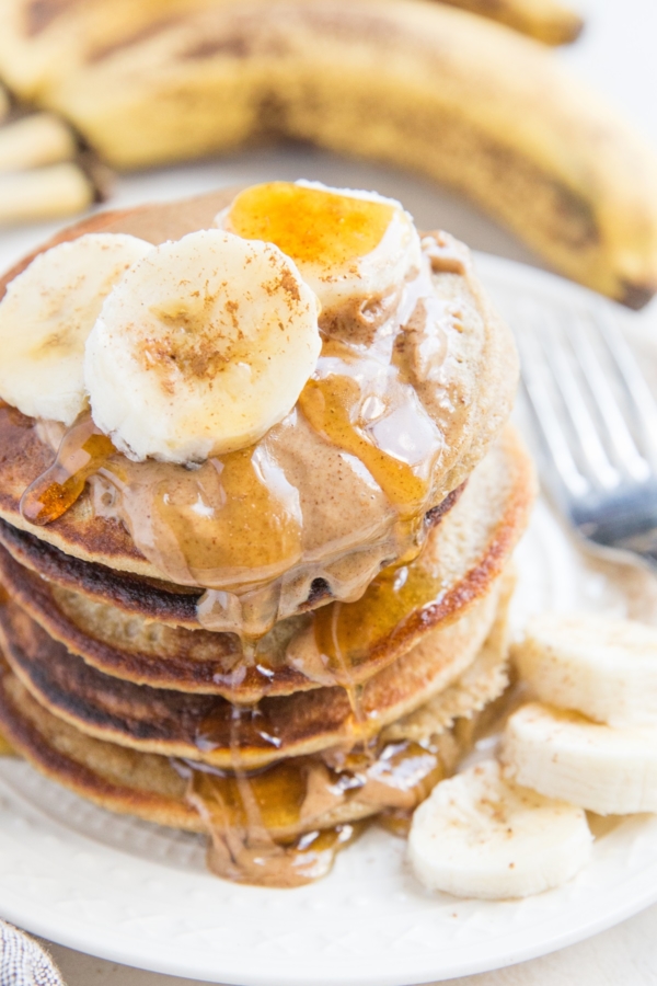 Stack of oatmeal banana pancakes on a plate with almond butter, honey, and sliced bananas on top.