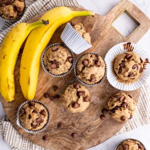 Gluten-Free Chocolate Chip Banana Muffins on a wooden cutting board with a napkin and fresh bananas to the side.