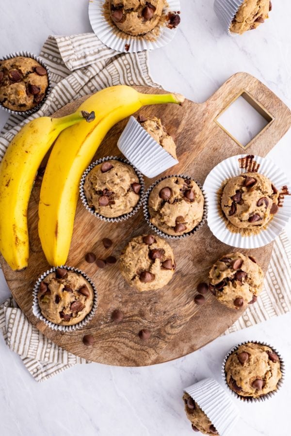 Gluten-Free Chocolate Chip Banana Muffins on a wooden cutting board with a napkin and fresh bananas to the side.