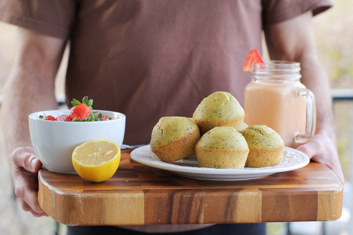 Man carrying a cutting board with a plate of Gluten Free Lemon Poppy Seed Muffins, a bowl of fruit, and a smoothie.