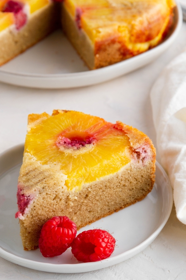 Slice of healthy pineapple upside down cake on a white plate with fresh raspberries to the side