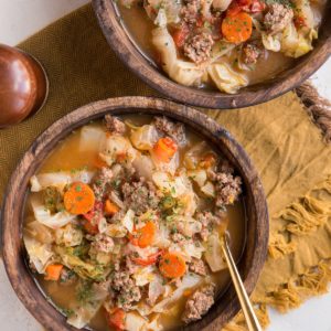 Two big wooden bowls of soup with a golden napkin and golden spoons on a white background.