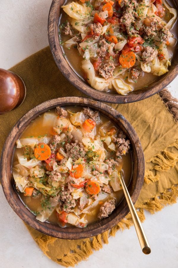 Two big wooden bowls of soup with a golden napkin and golden spoons on a white background.