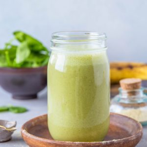 A green smoothie in a jar sitting on a wooden plate with ripe banana in the background. Ready to drink for a healthy meal replacement.