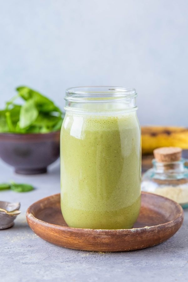 A green smoothie in a jar sitting on a wooden plate with ripe banana in the background. Ready to drink for a healthy meal replacement.