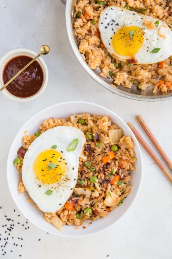 Two bowls full of kimchi fried rice with chopsticks and sesame seeds to the side.