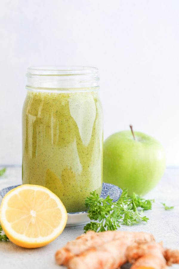 Large glass of smoothie with a green apple in the background, fresh parsley, a lemon and turmeric.