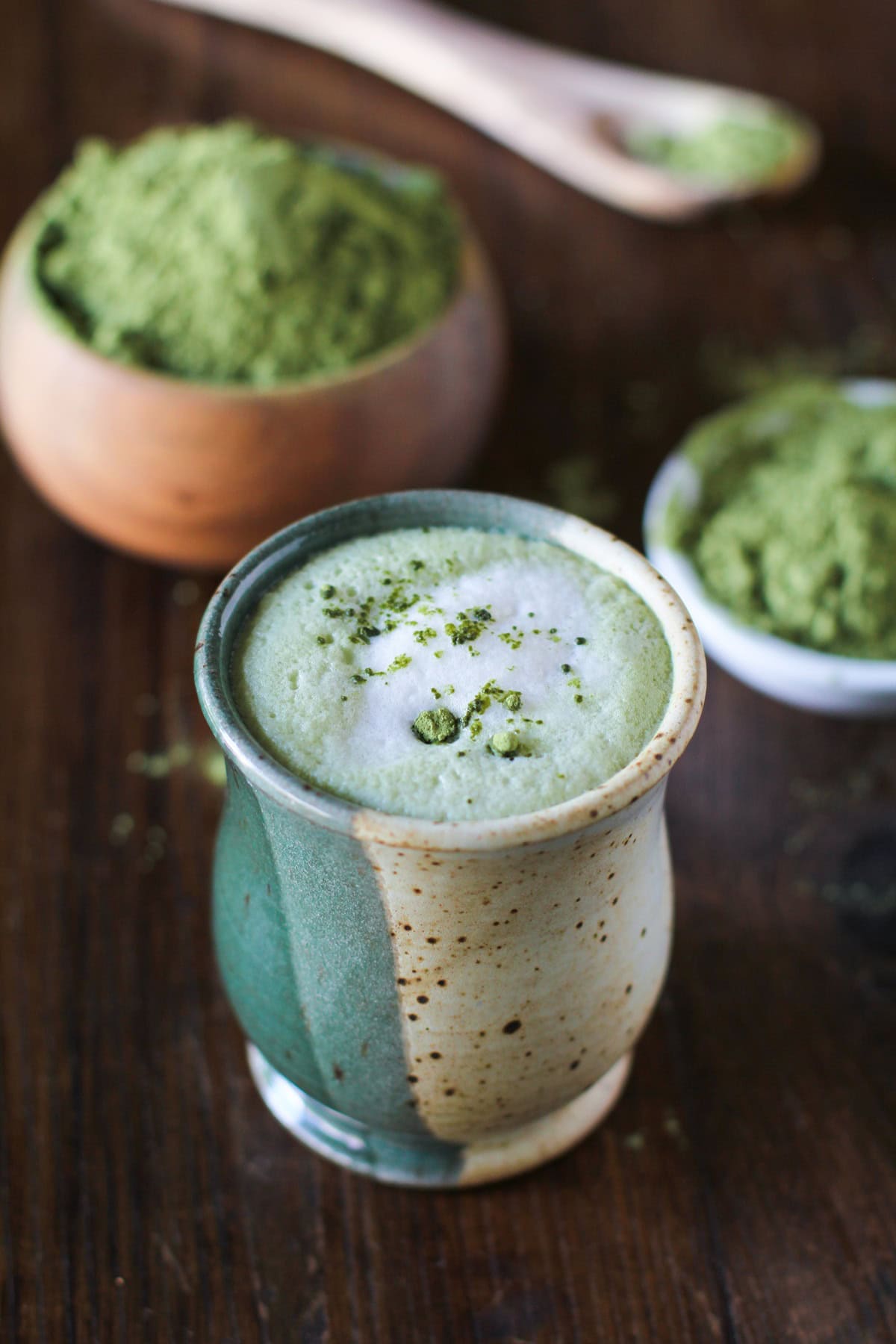 Ceramic mug full of matcha latte with a bowl of green tea matcha powder in the background