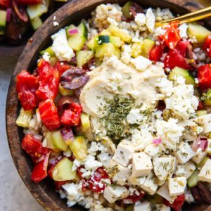 Wooden bowl full of chicken, rice, hummus, and cucumber tomato salad with a gold spoon and a bowl of salad to the side.